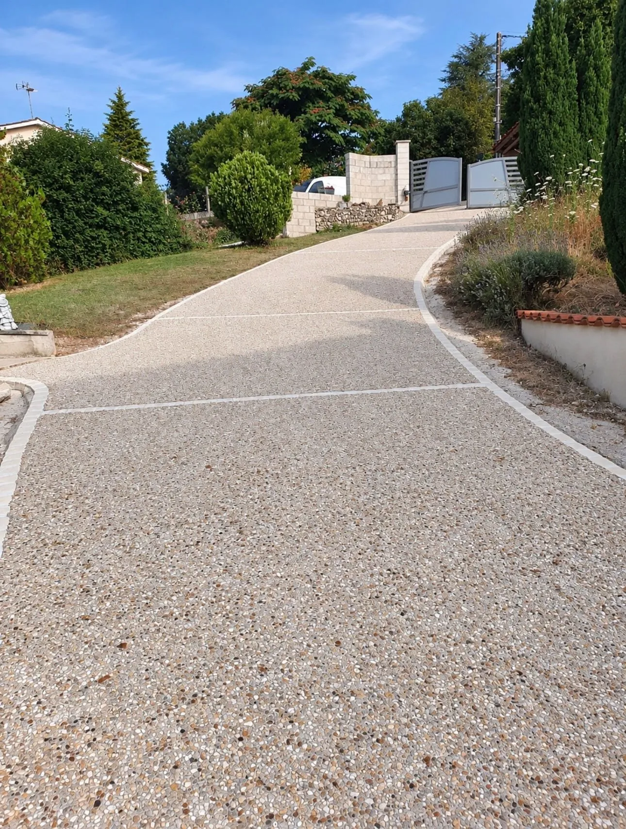 Allée en gravillons en pente, bordée de pavés clairs et menant à un portail