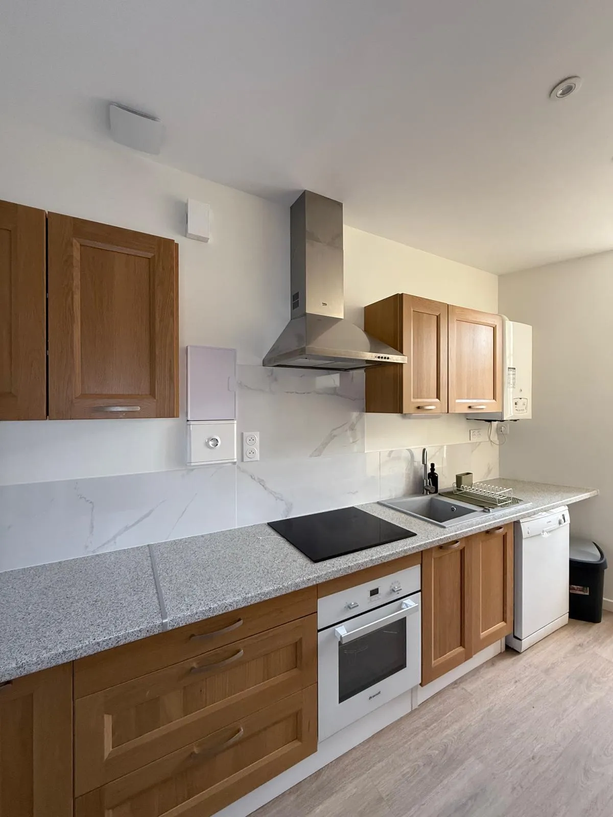 Cuisine aux meubles en bois, plan de travail granité gris, crédence effet marbre blanc, hotte inox et four encastré