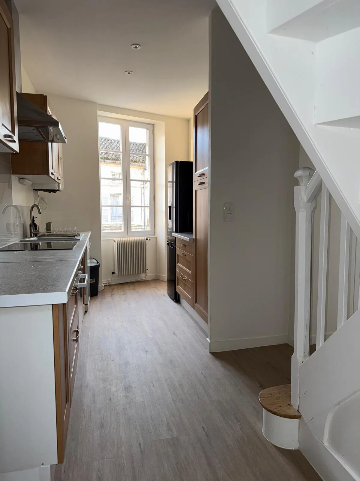 Cuisine en longueur aux meubles en bois, plan de travail gris et fenêtre au fond, escalier blanc à droite