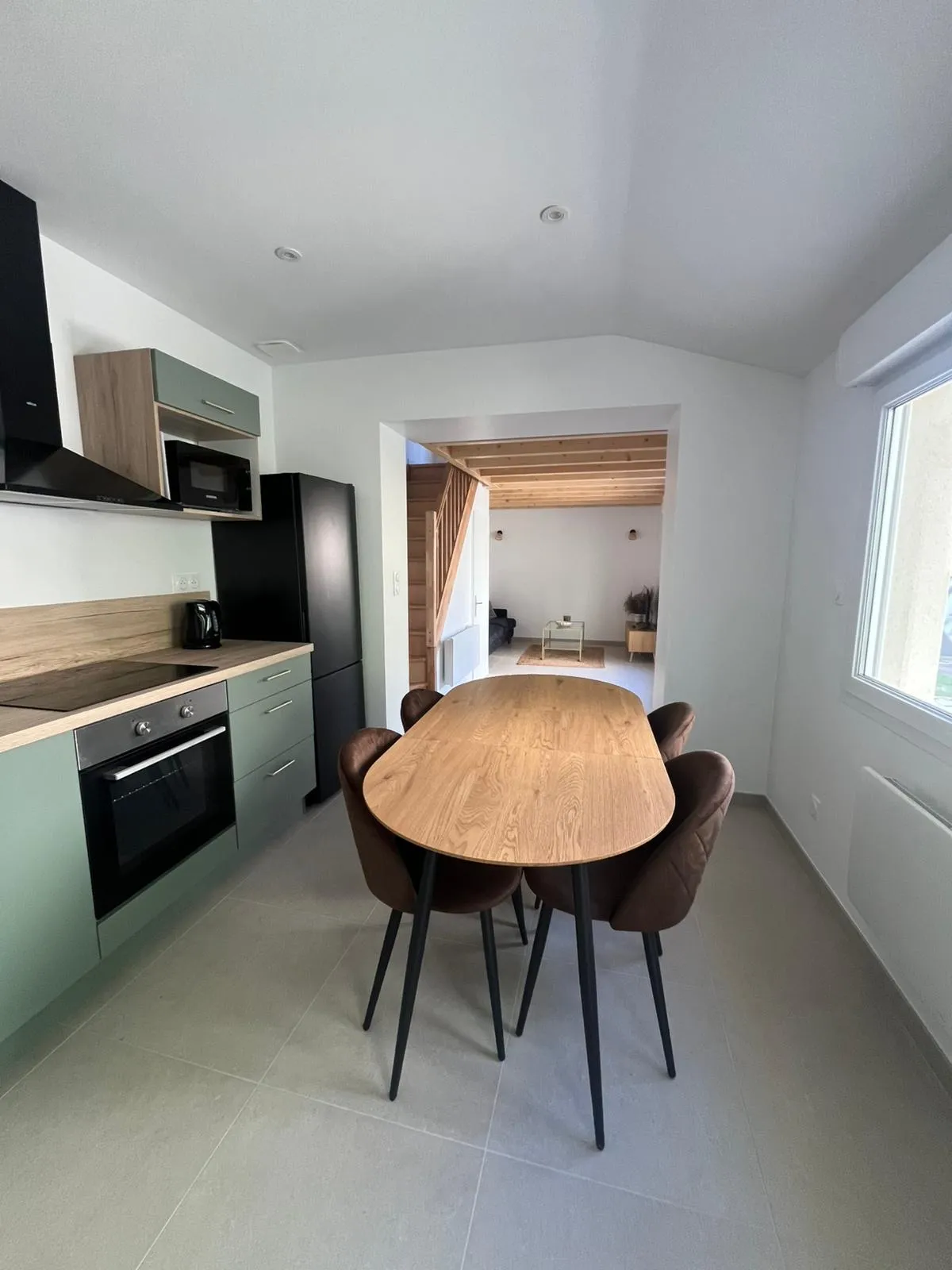Cuisine aux meubles vert sauge et plan de travail bois, ouverte sur un séjour avec escalier en bois