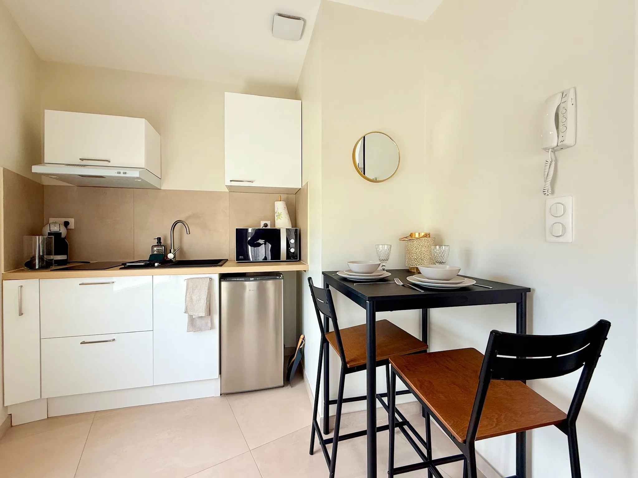 Kitchenette aux meubles blancs avec plan de travail bois clair, mini réfrigérateur et table haute avec deux tabourets