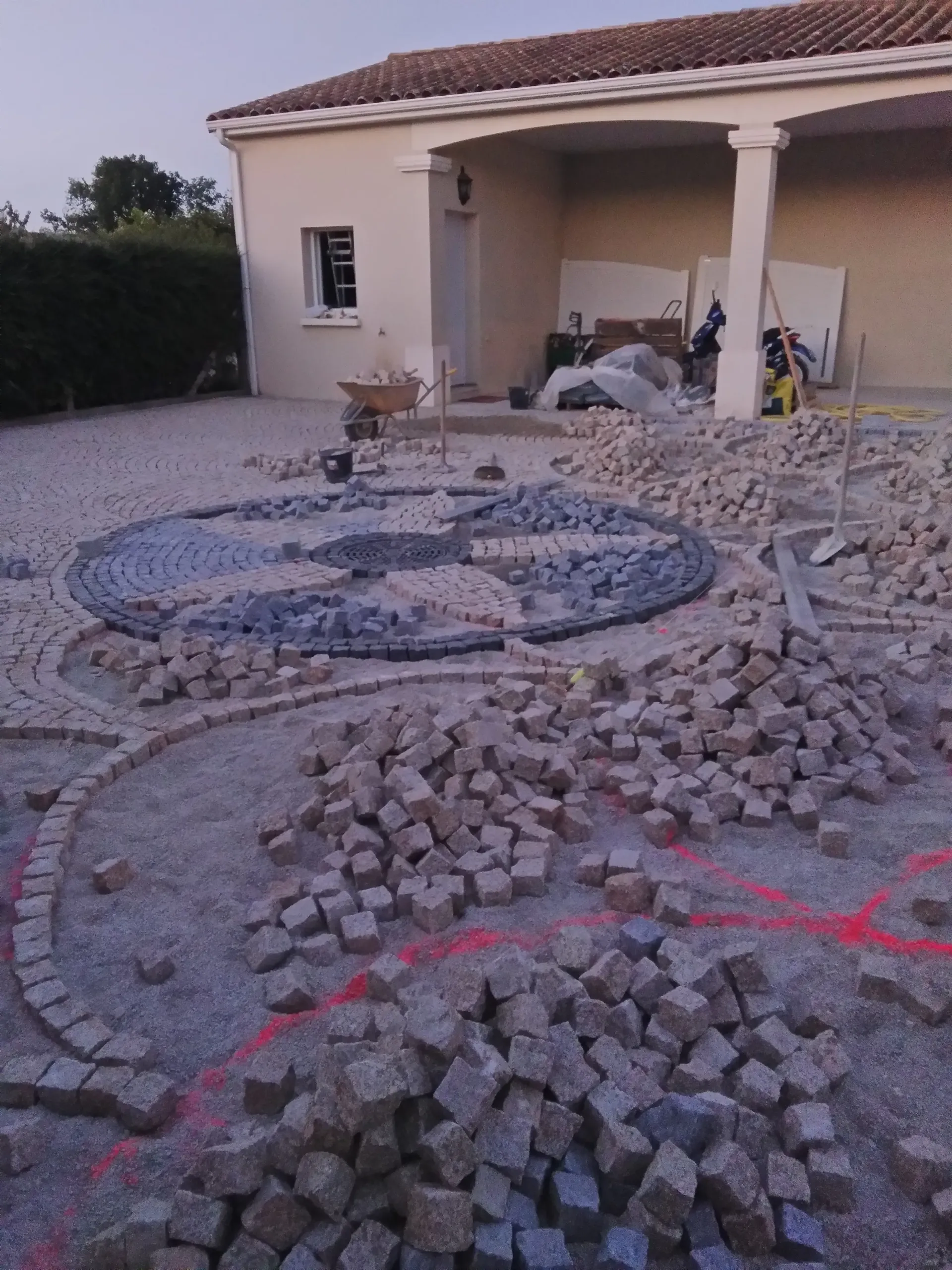 Chantier de pose de pavés en granit devant une maison, avec le motif circulaire en cours et des tas de pavés
