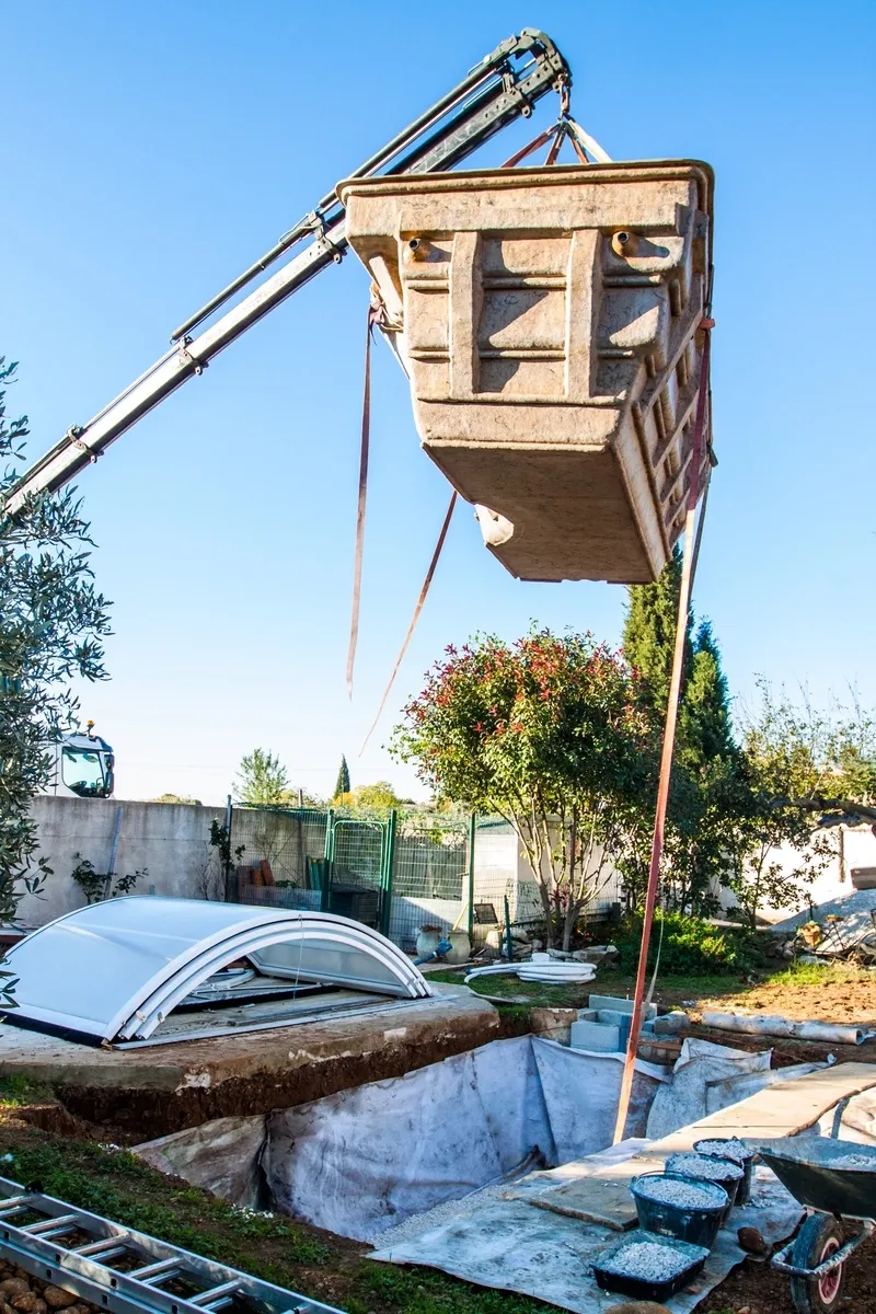 Coque de piscine suspendue à une grue au-dessus d’une fouille, sous un ciel bleu