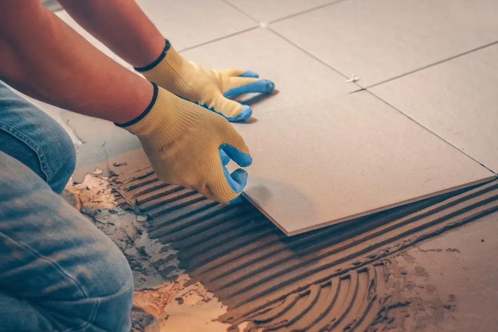 Gros plan de mains gantées posant un grand carreau de carrelage beige sur une colle crantée