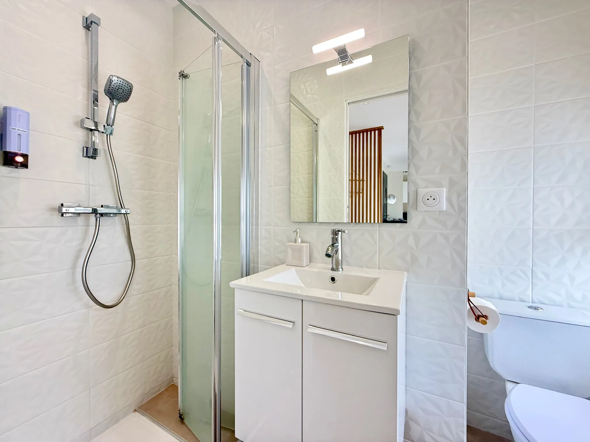 Salle d'eau aux carreaux blancs en relief, douche avec paroi vitrée et vasque sur meuble blanc