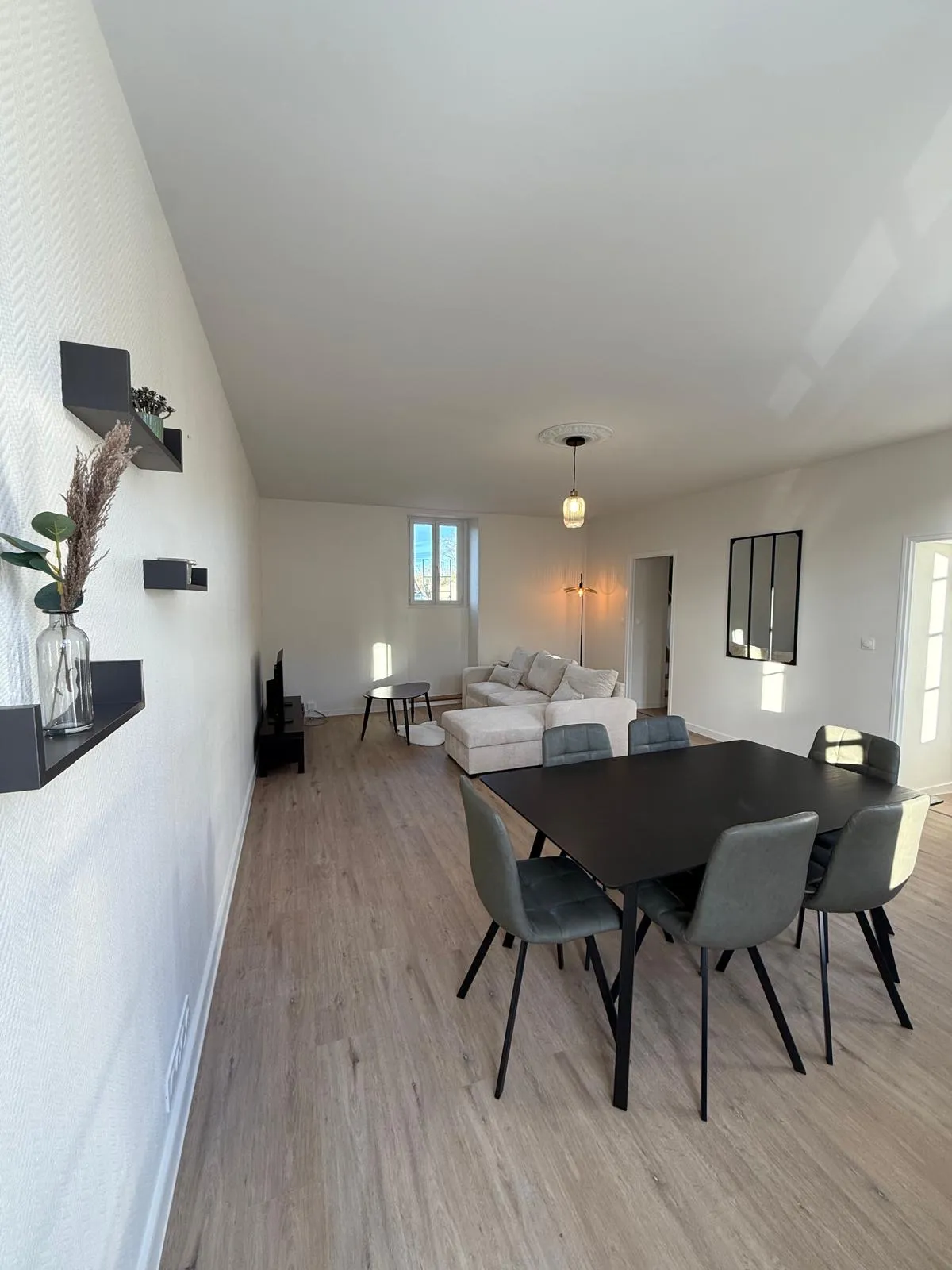 Grand séjour aux murs blancs, sol clair, table noire avec six chaises et canapé d'angle beige au fond